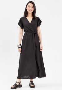 Robe noire portefeuille avec des manches courtes à volants, nouée à la taille, fabriquée en tissu texturé, associée à des sandales noires à lanières et des bracelets en argent.
