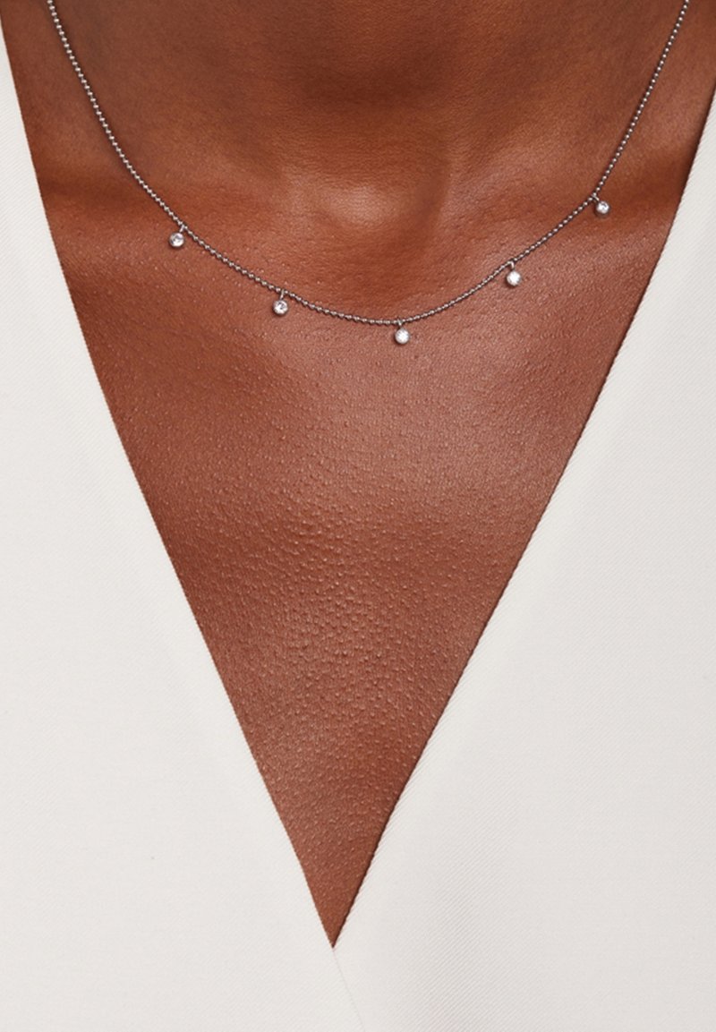 Collier chaîne argent avec cinq petits pendentifs ronds porté sur une peau foncée et un haut blanc à col en V.