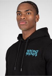 Sudadera negra con bolsillo tipo canguro, con un brillante logo azul que dice "WRONG FRIENDS". Incluye cordones y una textura de tela suave.