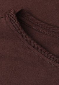 Nahaufnahme eines braunen Stoffes, der die Naht am Halsausschnitt und einen gefalteten Rand mit strukturiertem Besatz zeigt.