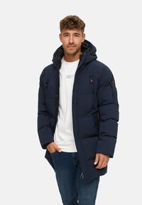 Chaqueta de pluma azul marino con capucha, que cuenta con múltiples bolsillos con cremallera y un diseño acolchado. Combinada con una camiseta blanca y jeans azules.