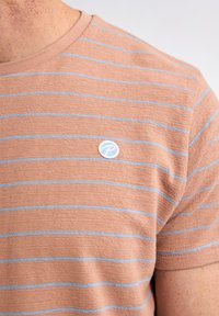 Hellbraunes kurzärmliges Hemd mit hellblauen horizontalen Streifen und einer strukturierten Stoffoberfläche. Verfügt über ein kleines kreisförmiges Logo auf der Brust.