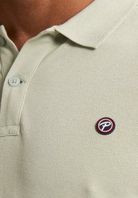 Polo shirt verde chiaro con tessuto texturizzato, dotato di colletto e due bottoni. Presenta una spilla con logo rotondo con accenti neri, rossi e bianchi.