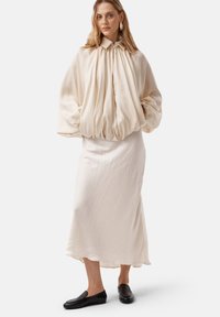 Crèmekleurige, textuur blouse met een hoge kraag en geplooid ontwerp, gecombineerd met een vloeiende, enkellange crèmekleurige rok en zwarte instapschoenen.