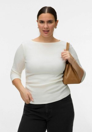 Kobieta o ciemnych włosach, ubrana w biały top i czarne spodnie, trzyma na ramieniu brązową torebkę, na jednolitym białym tle.