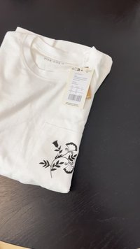 Hvid t-shirt lavet af bomuld med et sort blomsterdesign på venstre lomme. Etiket vedhæftet med plejeb instrukser og størrelsesoplysninger.