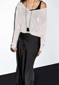 Vrouw draagt een licht gebreide trui met blote schouders over een zwarte top, een zwarte satijnen rok en een lange zwarte hangende ketting, tegen een witte achtergrond.