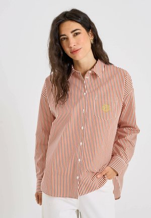 Femme aux longs cheveux foncés porte une chemise à rayures verticales marron et blanche avec des boutons et un petit logo soleil sur la poche poitrine, associée à un pantalon blanc.