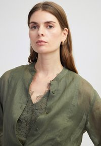 Olivegrünes, transparentes Hemd mit Knöpfen aus leichtem Material, mit Rundhalsausschnitt und darüber getragenem Spitzen-Tanktop.