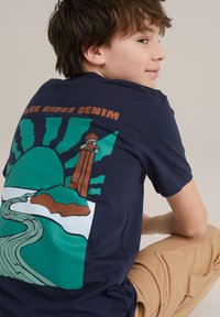 Navyblauwe T-shirt met een kleurrijke grafiek van een vuurtoren met groene en bruine accenten, ronde hals en korte mouwen, tegen een neutrale achtergrond.
