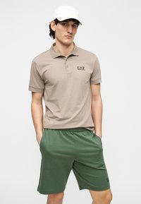 Beežist polosärk rinnal logoga, kombineeritud rohelistest lühikestest pükstest, millel on elastne vöökoht ja kaks küljetaskut. Peal valge nokamüts.