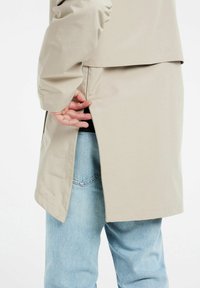 Cappotto lungo di beige chiaro con uno spacco sul retro, realizzato in materiale liscia. Presenta una vestibilità ampia e un design semplice. Indossato sopra dei jeans blu.