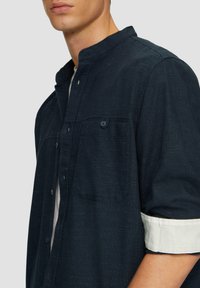 Chemise boutonnée bleu foncé en tissu texturé, dotée d'une poche poitrine unique et de poignets blancs contrastés retroussés.