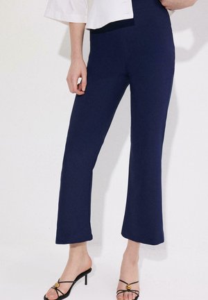 Kobieta nosząca granatowe spodnie 7/8 w połączeniu z czarnymi sandałami na obcasie ze paskami, stojąca na jednolitym białym tle.