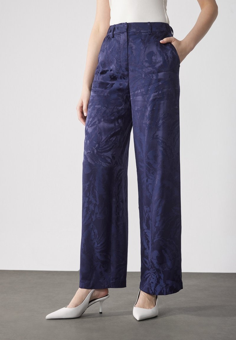 Femme portant un pantalon jacquard bleu foncé à taille haute et jambes larges avec motif floral, un haut blanc sans manches et des talons blancs à bout pointu.