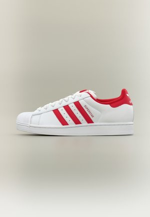 White sneaker with red accents, featuring three serrated stripes and a textured rubber toe cap. Smooth leather upper and padded collar.