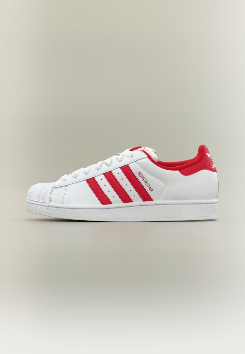 Fehér sportcipő piros részletekkel, három fogazott csíkkal és texturált gumi orrkappal. Simább bőr felsőrész és párnázott szegély.