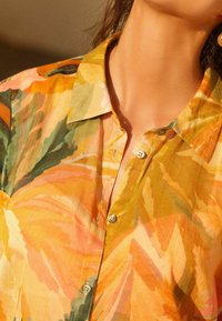 Camicia button-up floreale nei toni dell'arancione, verde e giallo. Caratterizzata da un colletto e un tessuto morbido con una texture semi-trasparente e bottoni decorativi.