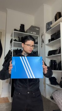 Caixa de cartão azul com três riscas brancas diagonais, segurada por uma pessoa vestida de negro, com botas visíveis ao fundo.