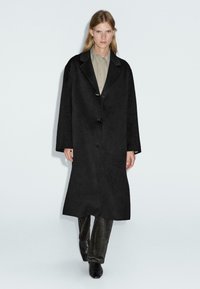 Cappotto di lana nera con vestibilità morbida, design a un petto, due grandi bottoni e profonde tasche laterali. Indossato sopra una camicia beige chiaro e pantaloni scuri.