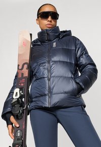Navy puffer jacket with a high collar and shiny texture; features a zip closure, side pockets, and is worn with black ski gear and skis.