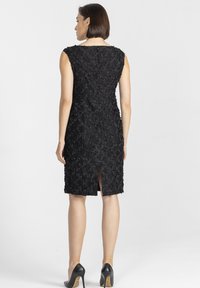Kobieta w czarnej, bezrękawnej, teksturowanej sukience do kolan z rozporkiem z tyłu oraz czarnych szpilkach, stojąca na jednolitym tle.