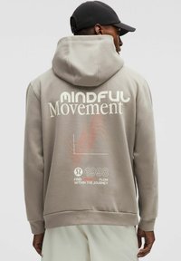 Hoodie gris avec une grande impression de texte blanc "MINDFUL Movement" dans le dos, présentant des graphismes de vagues rouges et l'année "1998" en bas.