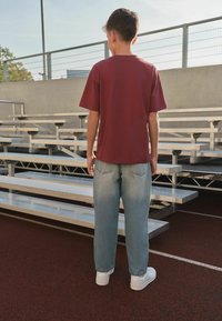 Junge trägt ein kastanienbraunes Hemd, hellblaue Jeans und weiße Turnschuhe, steht bei Tageslicht auf der Laufbahn und blickt auf leere Metalltribünen.