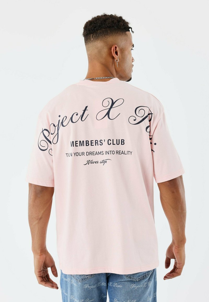 Hombre con cabello rizado corto, vistiendo una camiseta de gran tamaño color rosa claro con texto negro y jeans azules, de pie frente a un fondo blanco.