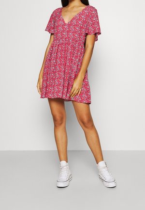 Vrouw die een korte rode bloemenjurk met wapperende mouwen en witte hoge sneakers draagt, staande tegen een effen grijze achtergrond.