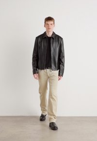 Svart skinnjakke med en glatt tekstur, kombinert med en svart skjorte, beige bukser og svarte sko. Modellen står mot en ensfarget bakgrunn.