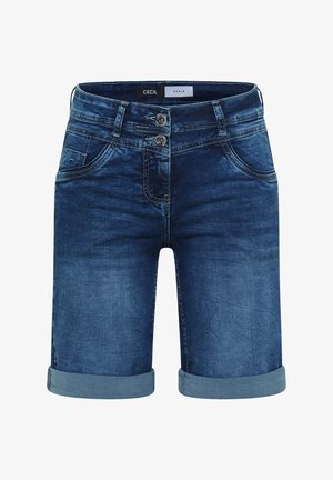 Frau trägt eine blaue Kappe, gestreiftes kurzärmeliges Shirt, aufgerollte Jeansshorts, beige Sneakers und hält eine große dunkelblaue Tragetasche.