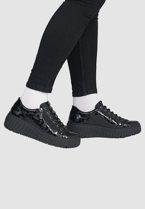 Person wearing shiny black platform sneakers with side zippers, white socks, and black fitted pants against a plain background.