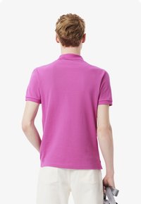 Rosa polo-shirt i bomull, med klassisk krage och korta ärmar, visad bakifrån med vita byxor och ett grått tillbehör i handen.