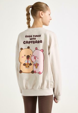 Person trägt einen cremefarbenen Pullover mit zwei Zeichentrick-Kapibaras, die Bubble Tea trinken, und dem Text "Gute Zeiten mit Kapibara" auf dem Rücken.