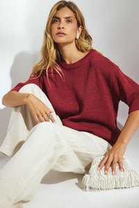 Czerwony, teksturowany sweter dziany z szerokimi rękawami, zestawiony z jasnobeżowymi spodniami lnianymi oraz białą torebką z frędzlami. Proste akcenty biżuteryjne.