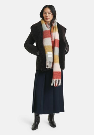 Frau in einer dunklen Schaffelljacke, einem langen, mehrfarbigen Streifen-Schal, navy-blauen, weit geschnittenen Hosen und schwarzen Stiefeletten, die mit den Händen in den Taschen steht.