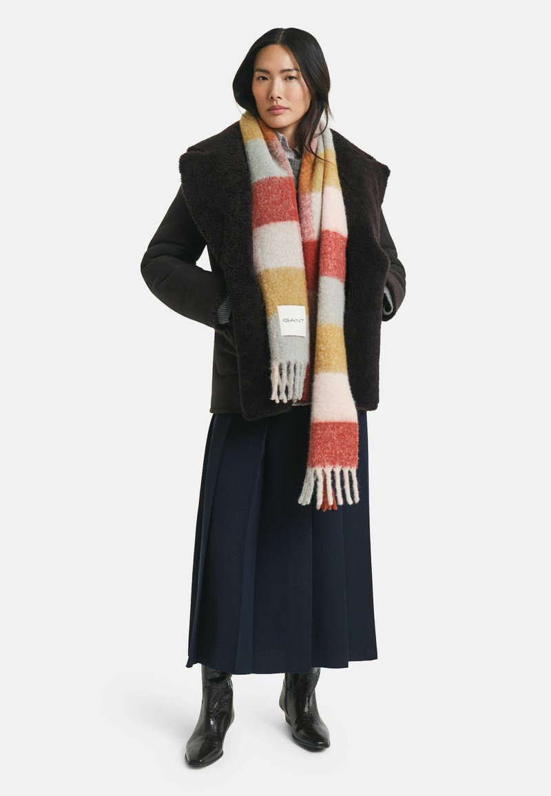 Femme portant une veste en shearling sombre, une longue écharpe rayée multicolore, un pantalon large bleu marine et des bottines noires, se tenant les mains dans les poches.