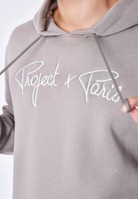Sudadera gris claro hecha de una tela suave, con una capucha amplia y el texto bordado "Project + Paris" en blanco. Los cordones son visibles.