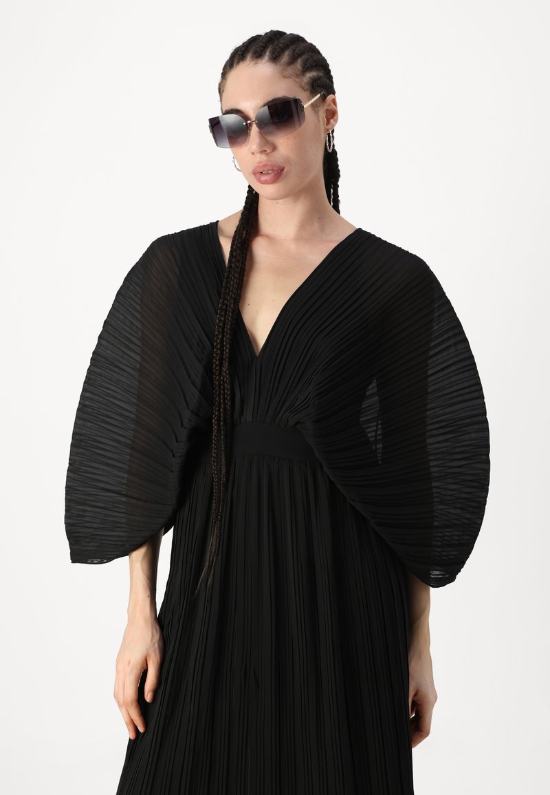 Schwarzes plissiertes Kleid mit dramatischen, durchsichtigen Ärmel-Details und einem tiefen V-Ausschnitt. Der Stoff ist leicht und hat eine weiche Textur.