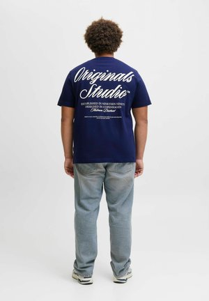 Marineblaue Baumwoll-T-Shirt mit weißem Text auf der Rückseite, versehen mit "Originals Studio" und zusätzlicher Markenkennzeichnung, kombiniert mit hellblauen Jeans.