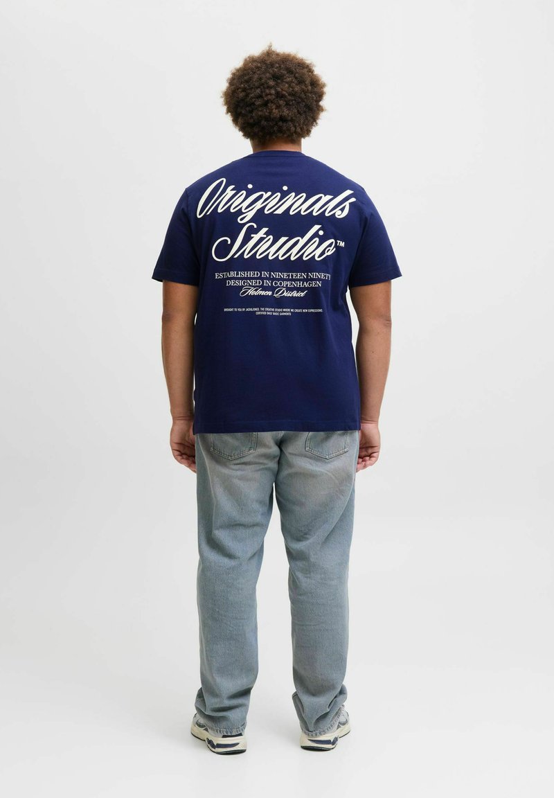 Mørkeblå bomullst-skjorte med hvit tekst på baksiden, med "Originals Studio" og ekstra merkevare, kombinert med lyseblå jeans.