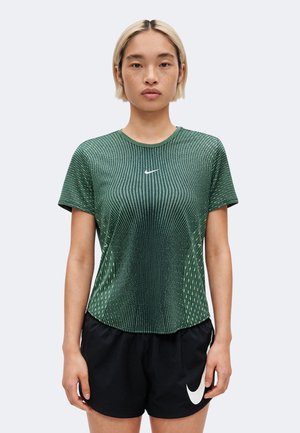 Jeune personne aux cheveux blonds courts portant un t-shirt vert à motifs Nike et un short noir Nike, debout devant un fond blanc.