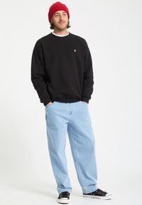 Sort sweatshirt, tasker lys blå jeans og sorte sneakers. Modellen bærer en rød strikket hue. Simpelt design med minimale accenter.