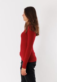 Maglione rosso a coste con maniche lunghe, forma aderente e scollatura rotonda. Indossato con pantaloni neri, esposto su uno sfondo bianco semplice.