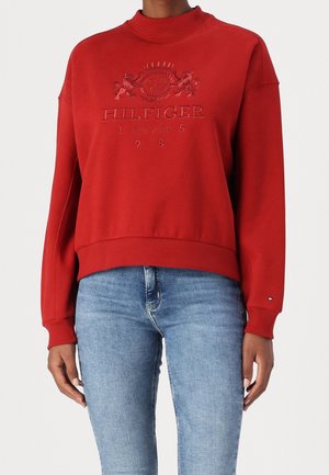 Personne portant un sweat-shirt rouge avec le logo en relief "Hilfiger New York 1985" et un jean bleu clair sur un fond uni.