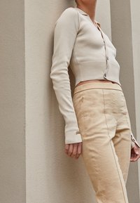 Personne portant un cardigan court beige et un pantalon taille haute de couleur beige clair, s'appuyant contre un mur texturé avec des rayures verticales.