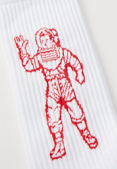 Chaussette blanche à côtes avec un design en tricot rouge représentant une personne en armure levant une main. Les détails notables incluent un tissu texturé et des couleurs vives.