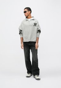 Sivi pulover oversized z ovratnikom in črnim logotipom, kombiniran s črnimi širokimi kavbojkami in sivimi ter belimi supergami. Gladek pleteni material.