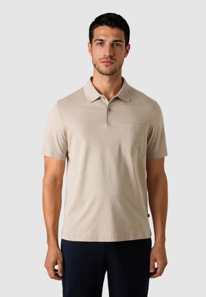 Mand iført en beige kortærmet polo med brystlomme og mørke bukser, stående foran en ensartet lys grå baggrund.